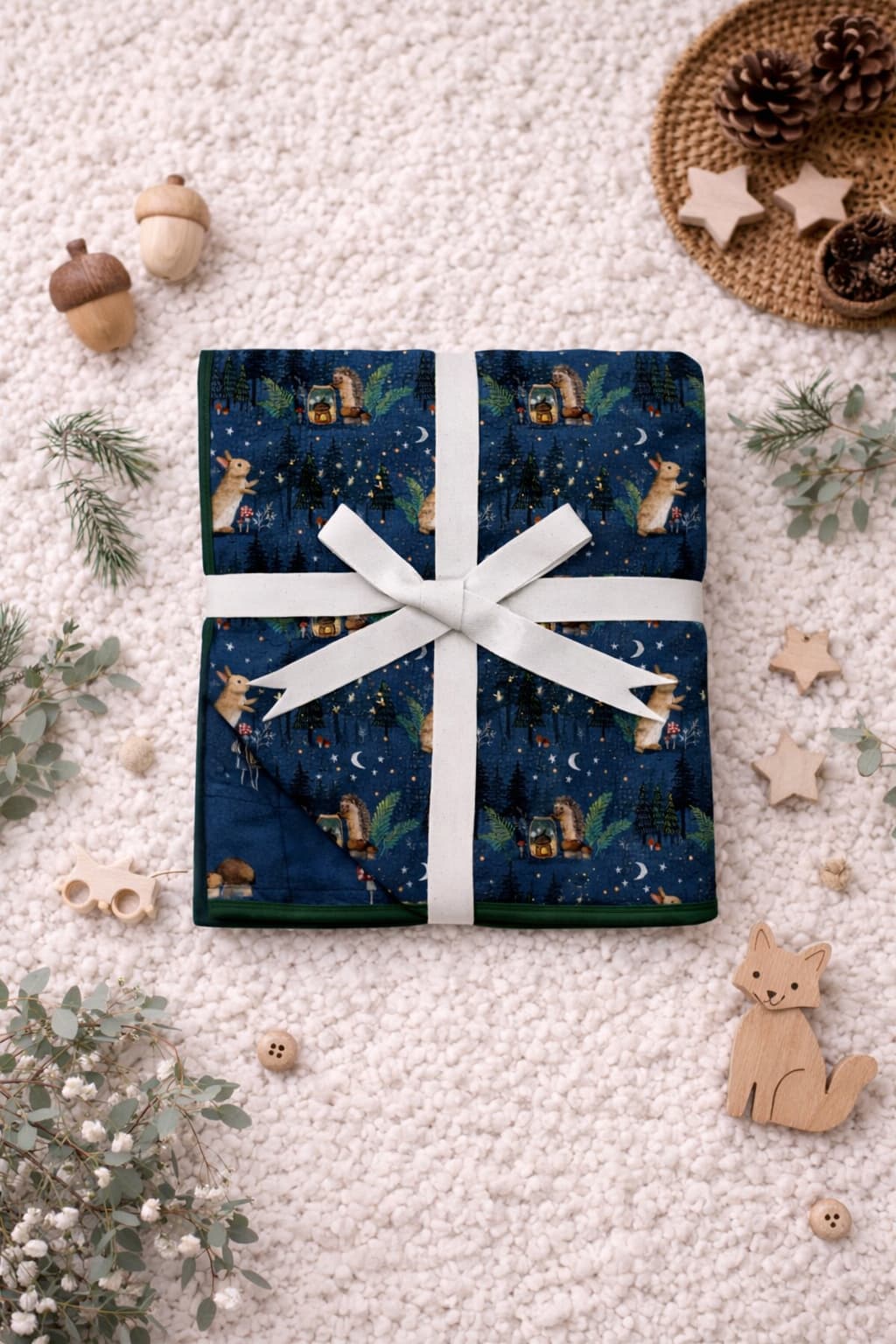 Bamboo blanket for toddler tied in a white ribbon on a soft surface. The bamboo blanket has woodland print showing hedgehogs, bunnies, fireflies, and more.